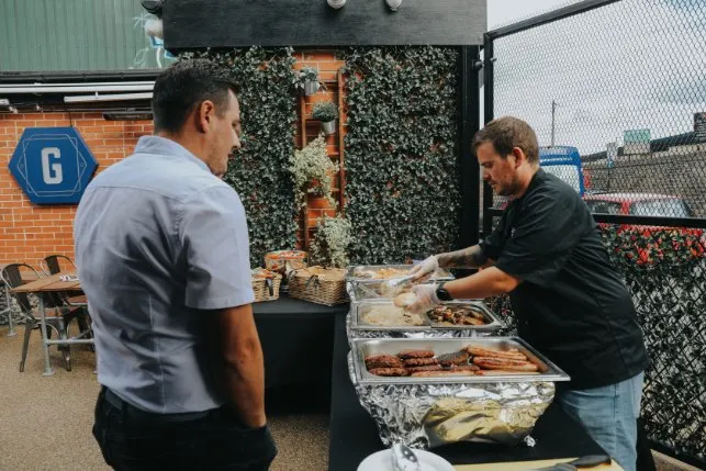 A chef serving sausages and grilled meat from foil trays at an outdoor buffet