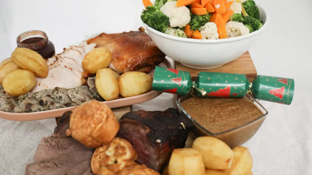 A Christmas dinner spread of sliced turkey and beef, roast potatoes, stuffing, Yorkshire puddings, gravy and a bowl of vegetables, with a cracker alongside