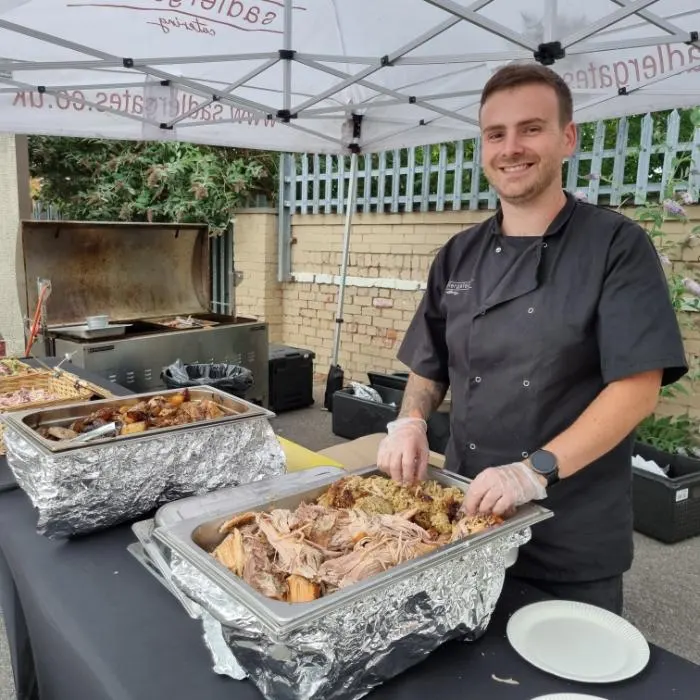 A Sadlergates chef serving pulled pork and stuffing from foil trays under a branded gazebo, with the hog roast machine behind