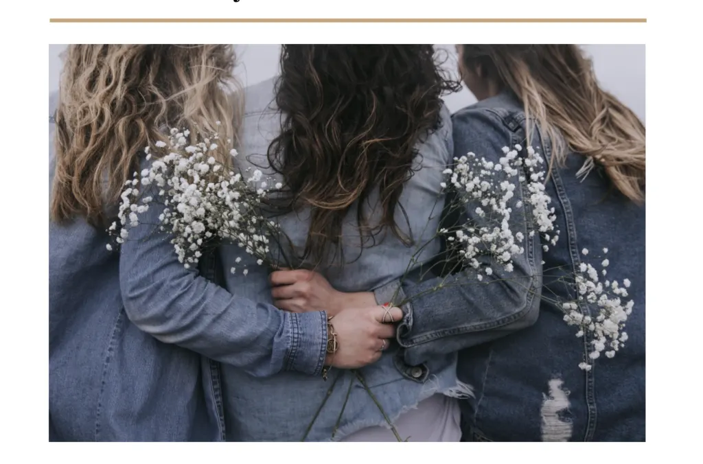 Three women photographed from behind in denim jackets, arms around each other, holding sprays of gypsophila