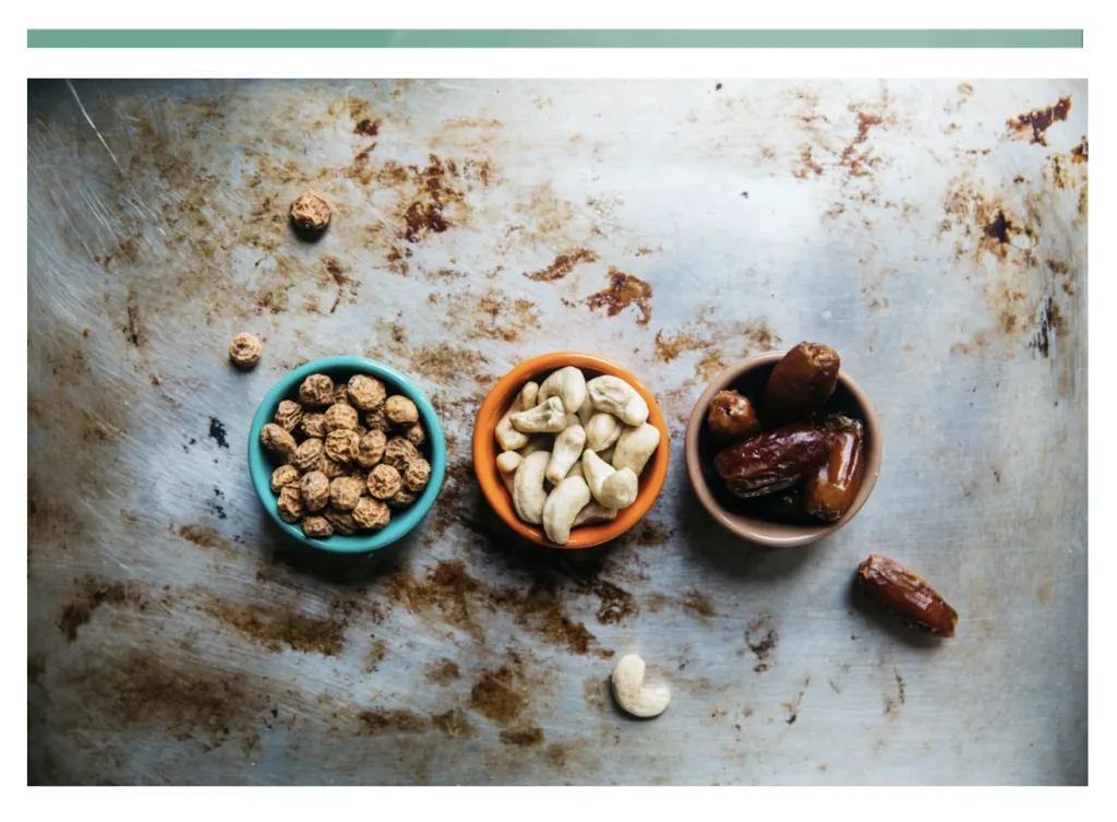 Three small bowls on a metal tray holding tiger nuts, cashews and dates