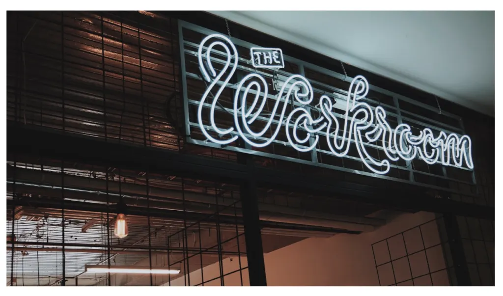 A white neon sign in cursive script reading The Workroom, above an industrial mesh partition