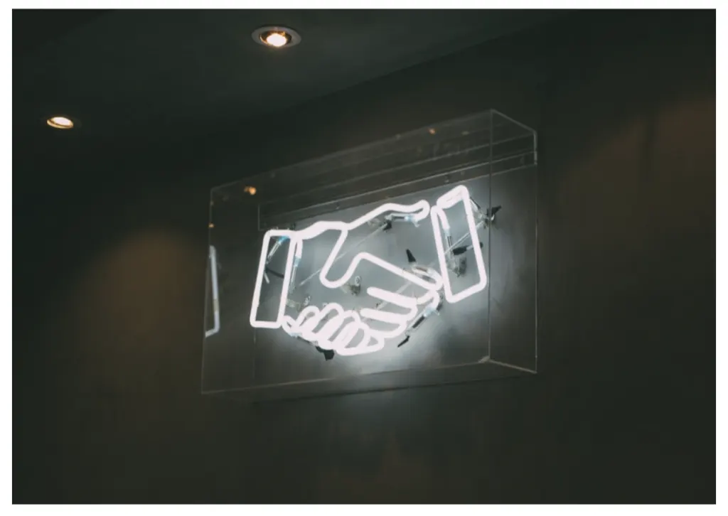 A white neon sign of two hands shaking, mounted in a case on a dark wall