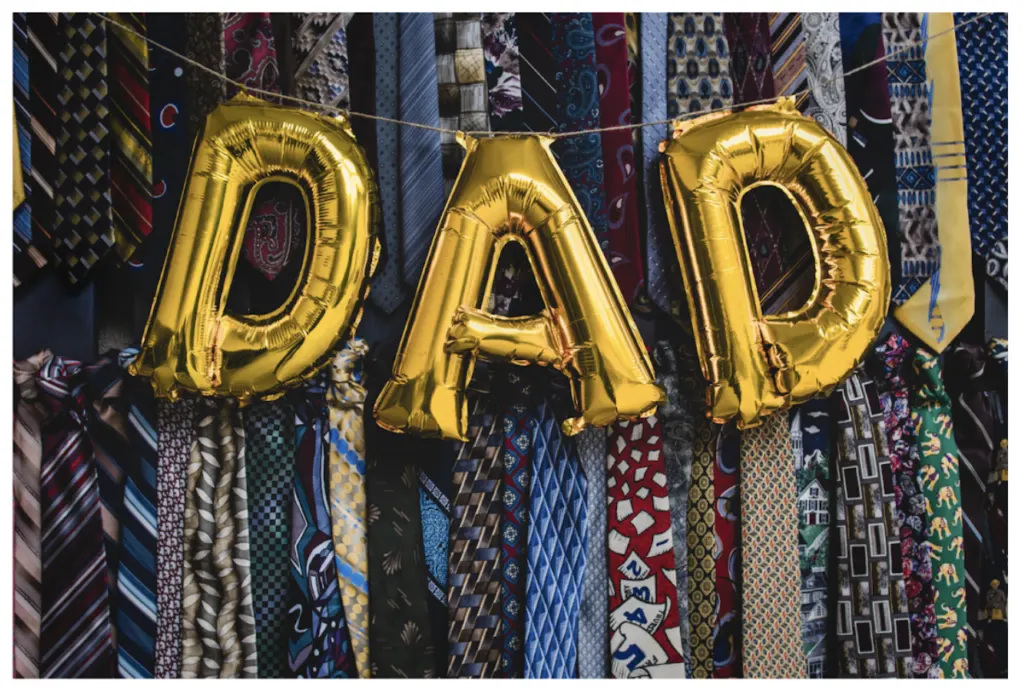 Gold foil balloons spelling DAD strung across a wall of hanging neckties
