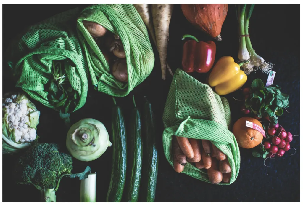 Vegetables photographed from above in reusable green mesh bags: carrots, potatoes, peppers, broccoli and cauliflower
