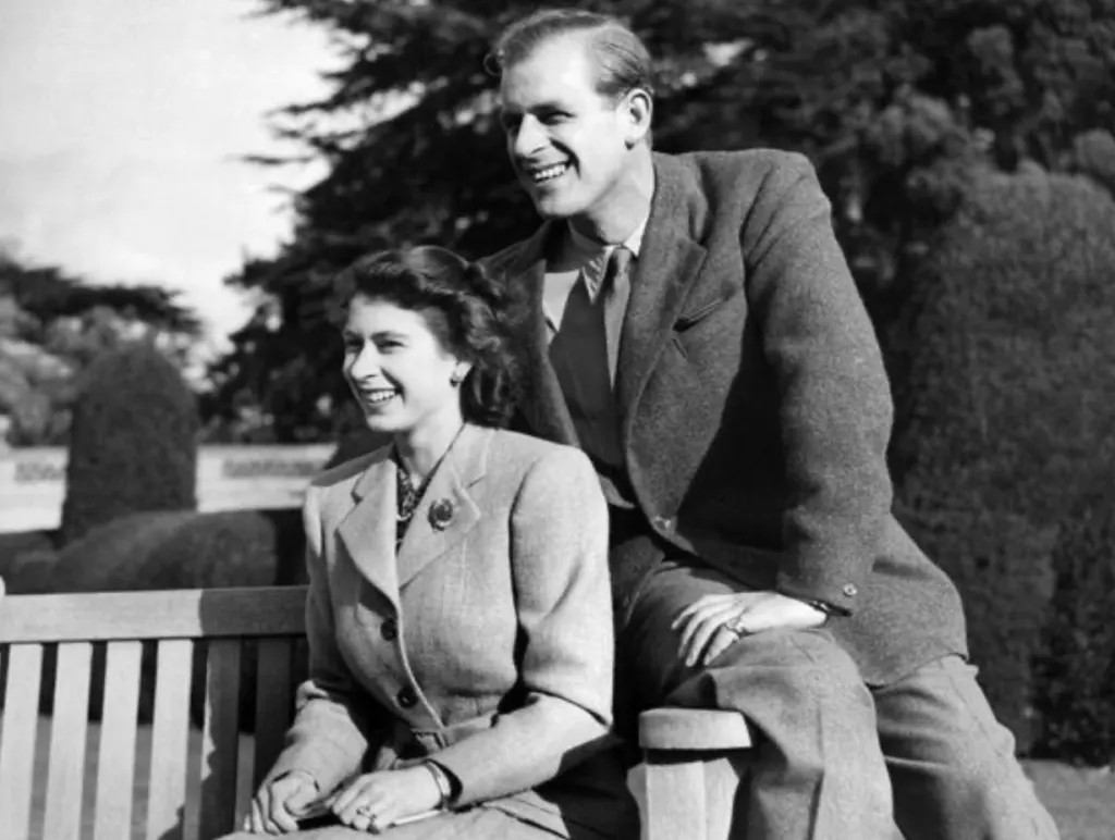 A black and white photograph of a young Queen Elizabeth II seated on a bench with the Duke of Edinburgh perched beside her, both laughing
