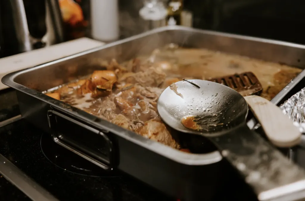 A roasting tray of meat in gravy on a hob, with a serving spoon resting across it