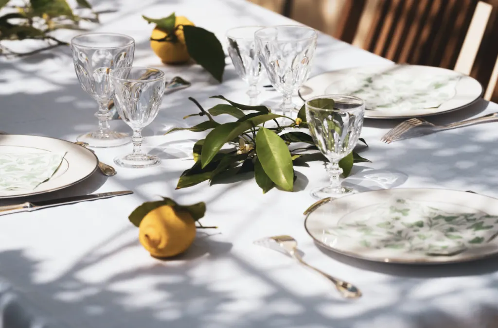 A table laid outdoors in dappled sunlight with cut-glass wine glasses, patterned plates, lemons and citrus foliage