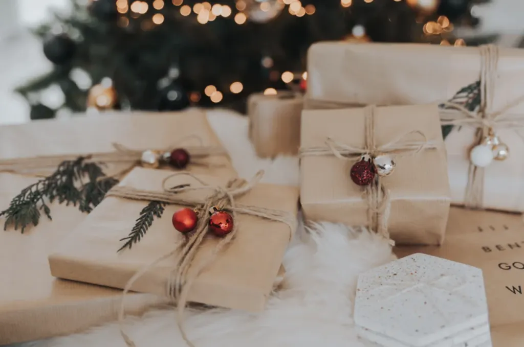 Christmas presents wrapped in brown paper and string, trimmed with red baubles and sprigs of foliage, in front of a lit tree