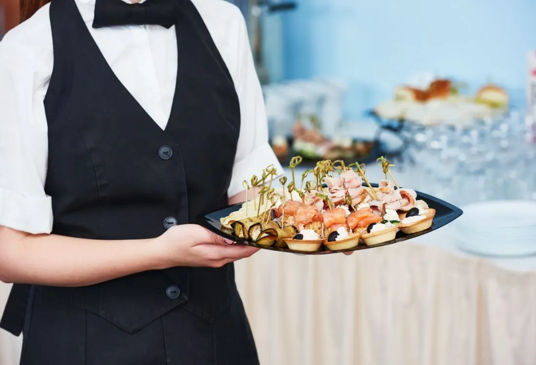 A waitress at a party handing out a platter of salmon and bruschetta created by the catering company who was hired to cater for the party for their variety.